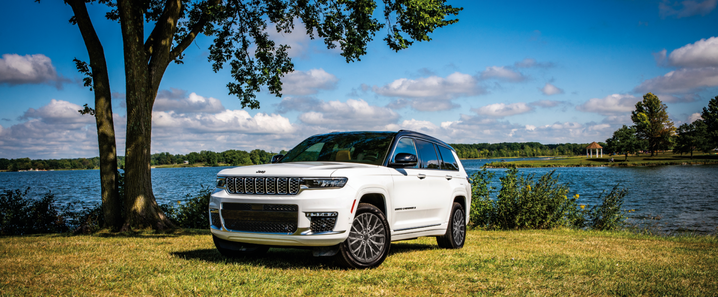 A gray 2024 Jeep Grand Cherokee Summit Reserve and a red Jeep Grand Cherokee Limited parked opposite of each other on a gravel mountain road at sunset.