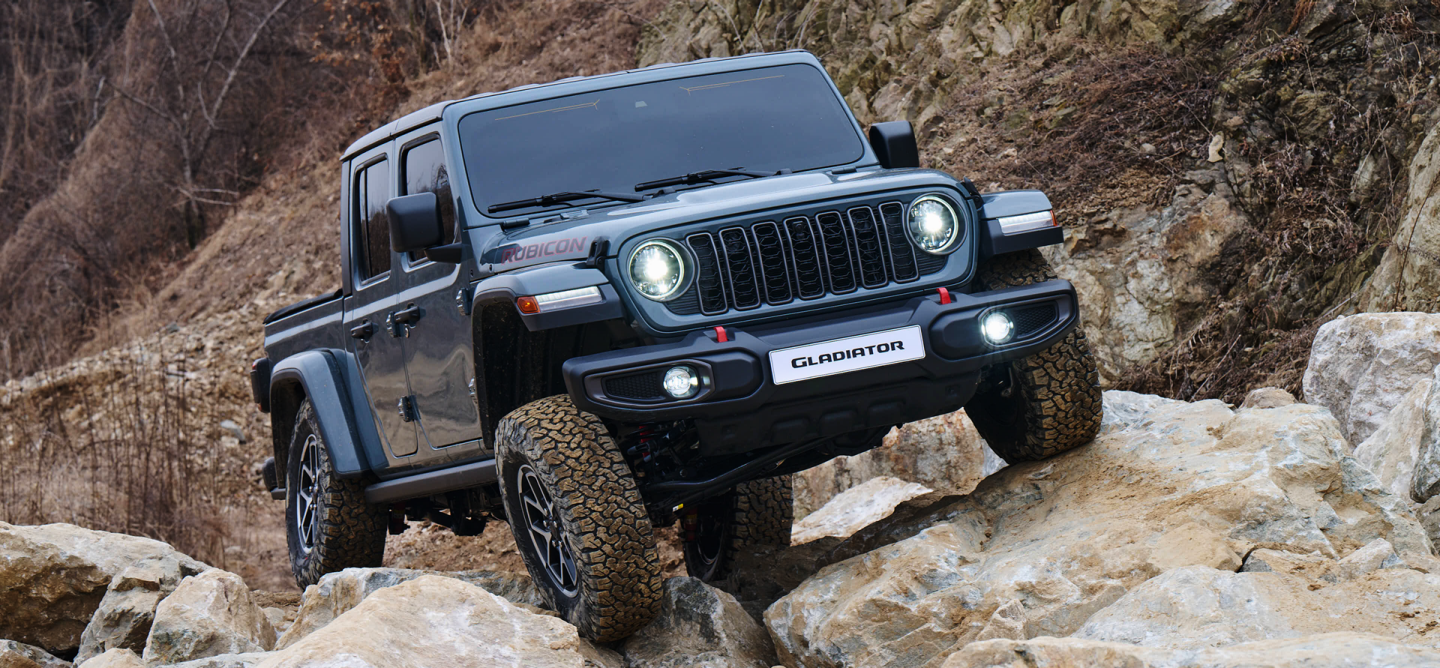 A blue 2025 Jeep Gladiator Mojave X with its doors and roof removed, ascending a trail in a hilly area off-road.