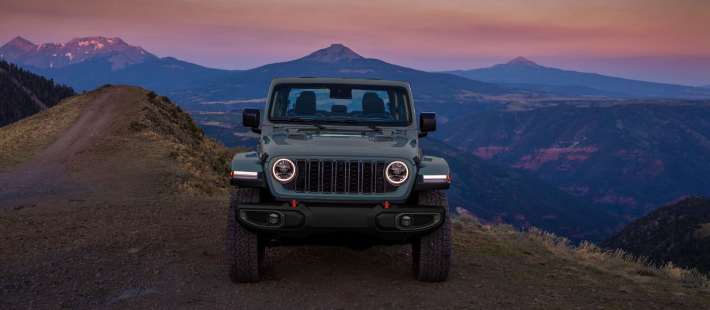 The front of a gray 2025 Jeep Gladiator Rubicon X parked on the edge of a cliff with mountains in the distance.