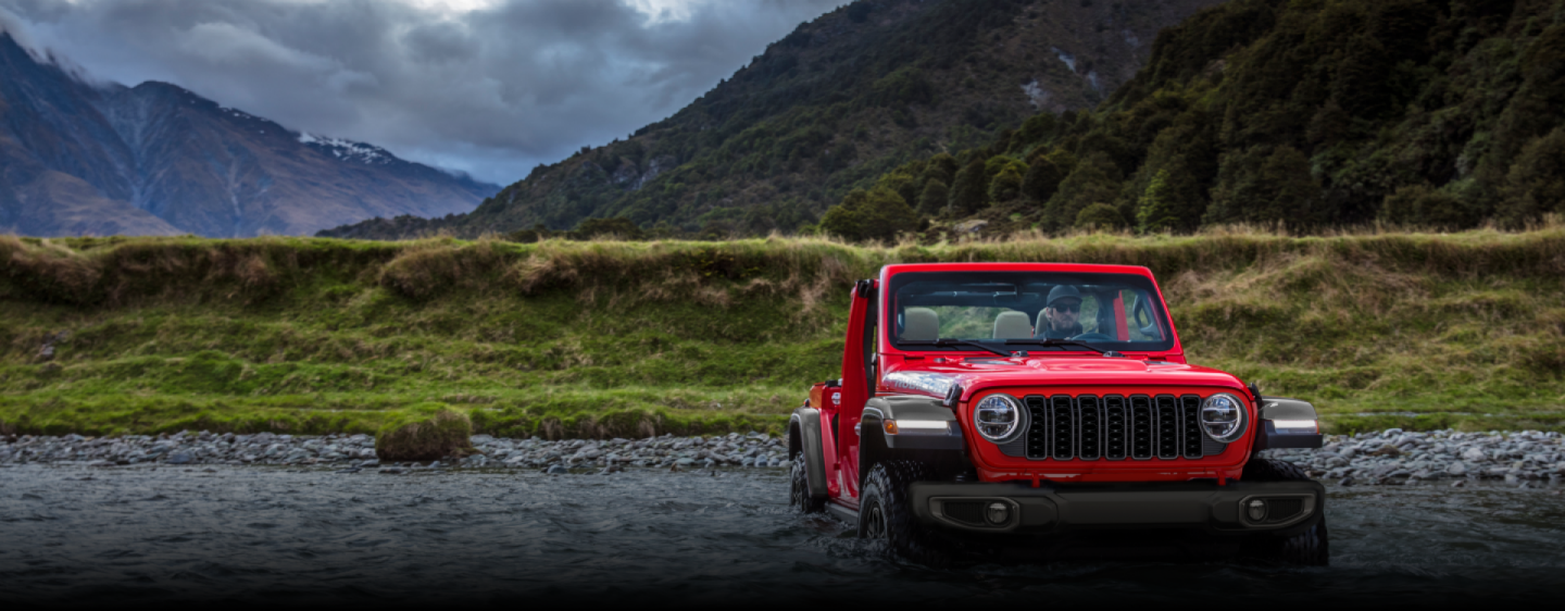 A front angle of a red 2024 Jeep Wrangler Rubicon being driven through a stream, following its path. Capability.