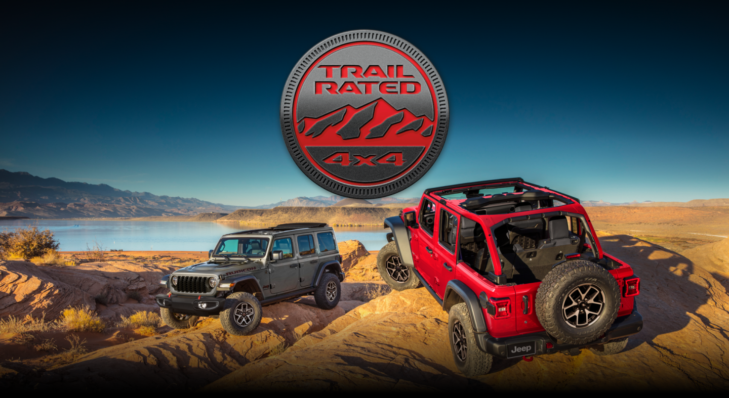 Two Jeep Wrangler models parked on sand filled with hills and ruts beside a lake. On the left, a silver 2024 Jeep Wrangler Rubicon 4xe. On the right, a red 2024 Jeep Wrangler Rubicon with its roof removed. Trail Rated 4x4.
