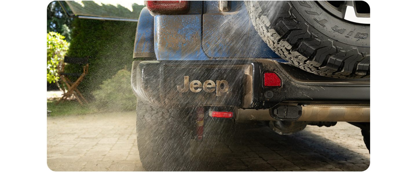 The license plate recess on the back of a blue 2026 Jeep Wrangler, with a hose spraying water on the vehicle, washing away mud. The corporate plate spells "Jeep".