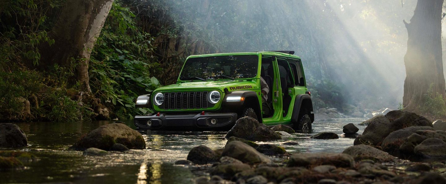 A lime green 2026 Jeep Wrangler Rubicon four-door, with its doors removed, fording through a stream in the woods off-road.