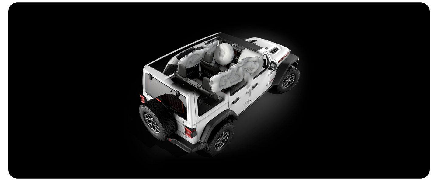 A blue 2026 Jeep Wrangler Rubicon X four-door, with its top removed and its headlamps on, parked on a rocky shoreline at the water's edge at night.