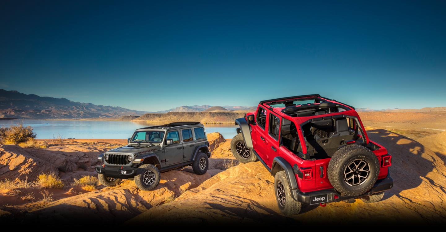 Two 2024 Jeep Wrangler models parked on a rocky clearing in the mountains. On the left, a gray Wrangler 4xe Rubicon four-door. On the right, a gray Wrangler Willys two-door.